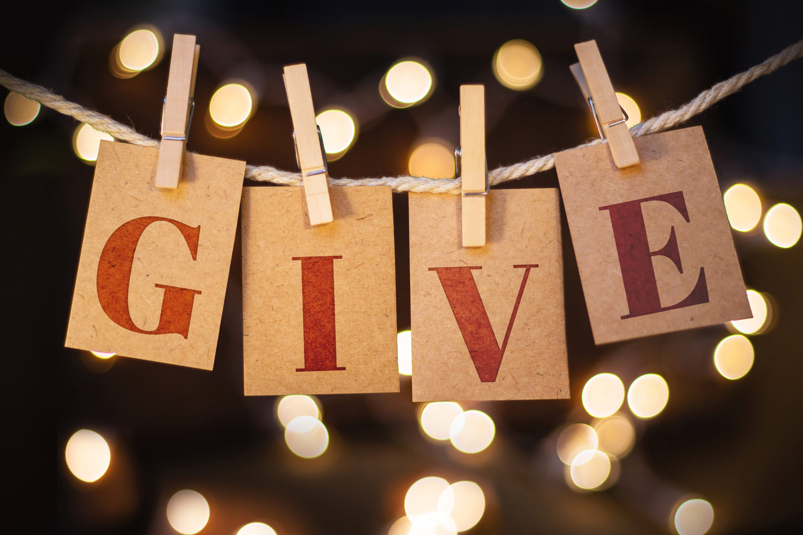 The word GIVE spelled out on clothespin clipped cards in front of glowing lights. Charitable Giving and Financial Planning.